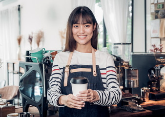 Fototapeta na wymiar asian woman barista wear jean apron holding hot take away coffee cup served to customer with smiling face at bar counter.cafe restaurant service concept.waitress working.
