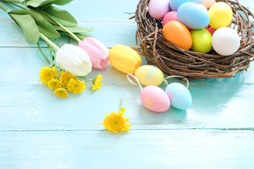 Colorful Easter eggs in nest with flowers on blue wooden background.  Easter holiday in spring season, top view with composition.