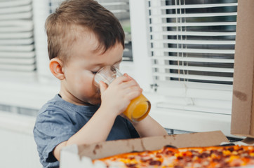 the child in the kitchen drinks orange juice and there is a large pizza