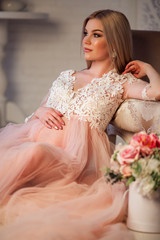 Naklejka premium Beautiful pregnant young woman is wearing luxury pink lingerie dress at home in cozy interior with bouquet of flowers