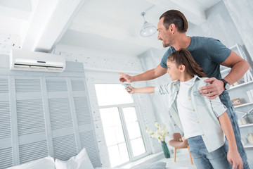 Comfortable temperature. Positive happy joyful man standing near his daughter and pointing at the air conditioner while teaching her how to use it