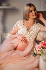 Naklejka premium Pretty blonde pregnant young woman is wearing luxury pink lingerie dress at home in cozy interior with bouquet of roses flowers