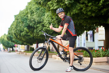 Fototapeta premium Happy man cyclist in professional cycling clothing and protective garment showing thumbs up sitting on bicycle in front of green trees and smiling to camera. Sportsman satisfied after morning training