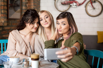 Obraz premium Female Friends In Cafe Taking Selfie Using Smart Phone. Women having fun, Smiling Drinking coffee and enjoying their time.