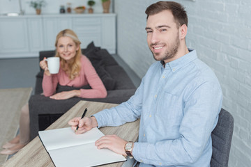 selective focus of smiling man at table with notebook and mother with cup of coffee at home