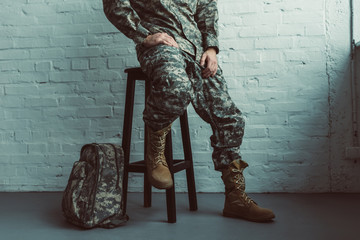 partial view of soldier in military uniform sitting on chair against white brick wall - Powered by Adobe