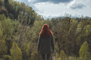 Longhair redhead girl wearing a grey coat standing on the edge of the hill in front of the forest...