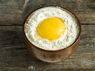 Wooden bowl with flour and broken chicken egg on rustic background