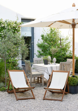 Gravel terrace style townhouse, with wooden deckchair, umbrella in white fabric, table covered with linen and plants in pots
