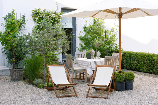 Gravel terrace style townhouse, with wooden deckchair, umbrella in white fabric, table covered with linen and plants in pots