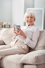 Pleasant morning. Pleasant senior woman sitting on the couch and posing for the camera while holding a cup of tea