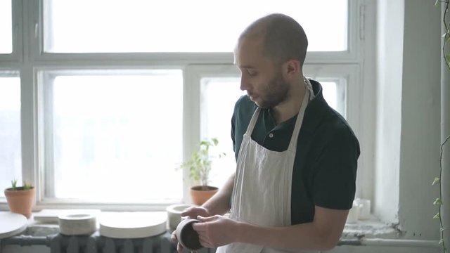 Portrait of ceramist, who is finishing his work on mug in bright studio. Young man with beard and in white dickey is taking the cup away from throwing wheel, standing up and doing out with the big