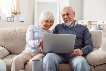 Quality time. Upbeat senior husband and wife holding a laptop in their lap and surfing the Internet together while sitting on the sofa in their living room