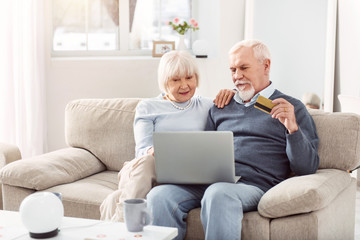 Convenient service. Upbeat elderly couple sitting on the couch and using their bank card while paying for public utilities online