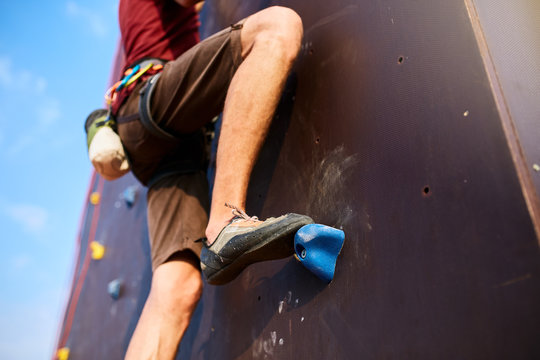 Bottom close up view of rock climber foot on training artificial climbing wall outdoors. Man feet in climbing shoes on rocks with equipment - belaying harness, magnesium chalk and carabiners.
