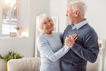 Enjoy dancing. Happy charming elderly woman smiling at the camera while dancing waltz with her husband in the living room