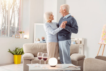 Together forever. Happy elderly couple dancing waltz together in the living room and looking at...
