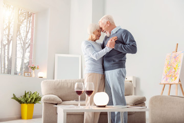 Deep in love. Pleasant elderly couple dancing waltz in the living room and leaning their foreheads against one another while smiling at each other