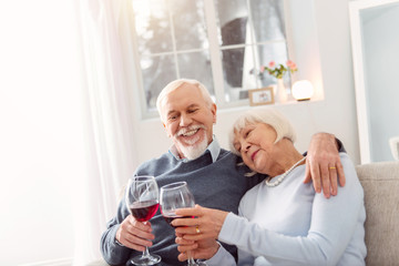 Relaxing together. Cheerful elderly couple sitting on the couch and cuddling while celebrating their wedding anniversary and drinking wine