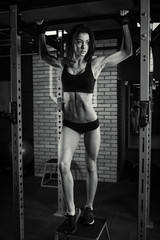 Fototapeta na wymiar Fit girl with black hair wearing black short top, shorts and gloves standing with horizontal bar on a box in gym, brick wall at background. Black and white portrait.