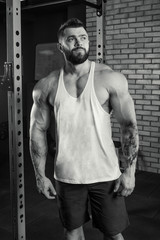 Muscular man with tattoos and beard standing in a white tank top and blue shorts in the gym. Black and white portrait.