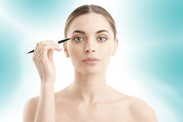 Studio shot of a beautiful young woman applying makeup to her eyebrow while standing at isolated background.