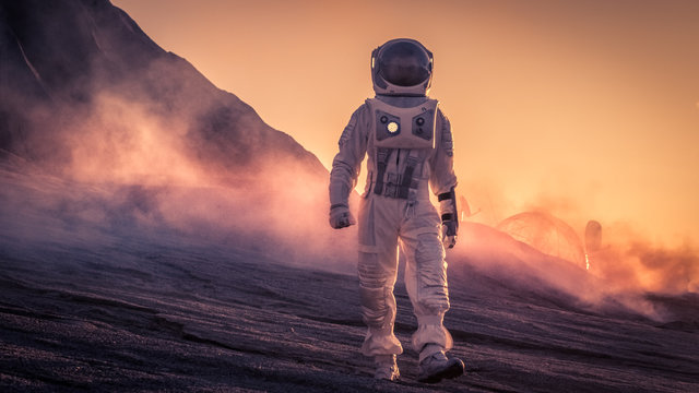 Astronaut Wearing Space Suit Walks on the Red Planet/Venus During Sunset. In the Background His Base with Rover Parked, Hot Red Daylight Sun Shines.