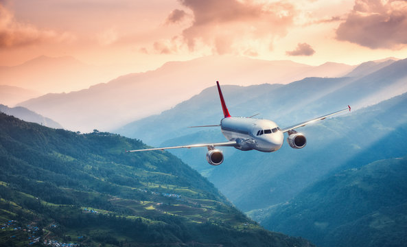 Fototapeta Aircraft is flying over green hills against mountains with yellow sunbeams at sunset. Landscape with passenger airplane, colorful sky, village. Passenger aircraft. Business travel. Commercial plane