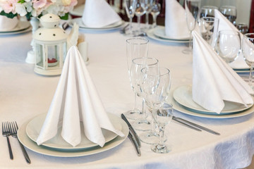 Glasses, flowers, fork, knife, napkin folded in a pyramid, served for dinner in restaurant with cozy interior. Wedding decorations and items for food, arranged by the catering service on a large table