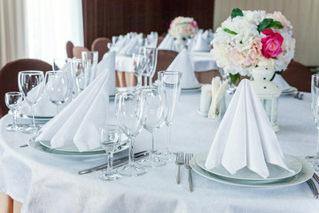 Glasses, flowers, fork, knife, napkin folded in a pyramid, served for dinner in restaurant with cozy interior. Wedding decorations and items for food, arranged by the catering service on a large table