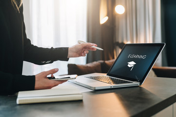Woman standing near table,pointing pencil on laptop with inscription on screen e-learning, image of square academic cap.