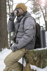 Obraz premium Handsome, bearded man in glasses with a camera and a backpack sitting on a tree stump in the winter forest and is holding a cup with a hot drink from a thermos.