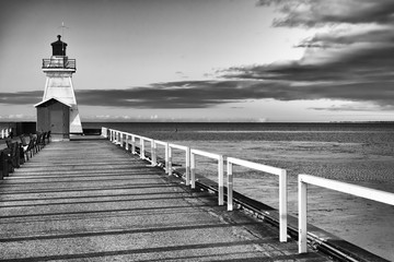 Old fashioned lighthouse on the end of a pier, vast water and cloud beyond. Black and white art