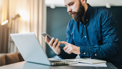Smiling bearded businessman sitting at table in front of computer, using smartphone.Freelancer works home, distance work