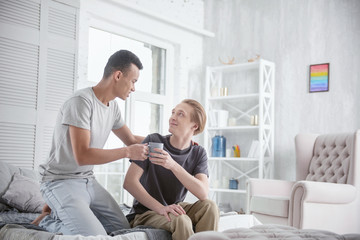 Sweet moments. Charming energetic gay couple posing on bed while gazing at each other and holding cup