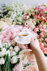 Woman holding delicious freshly brewed morning coffee in a white cup with a saucer on a flower bed background at the florist shop