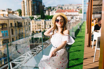 Funny girl with long hair  in sunglasses is moving around on the terrace in cafe . She wears a white dress with bare shoulders, red lipstick  and hat . She looks happy.