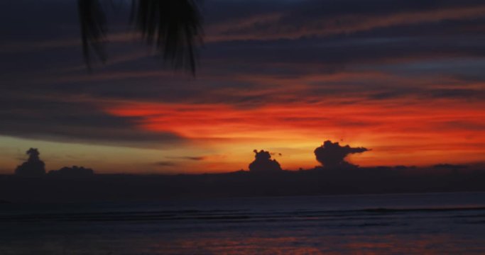 Red, orange, blue colors in the sky among clouds, sea or ocean water, palm leaf like in paradise