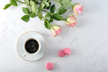 A cup of coffee and a popular makarun of pink color. There are several roses in the frame. Light marble background. View from above.