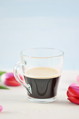 Transparent cup with coffee surrounded by pink tulips and pink macaroons. Light background. Close-up. Macro photography. Vertical orientation of the frame.