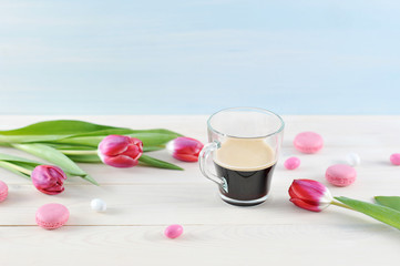 Pink tulips and pink macaroons. The composition is complemented by a transparent cup of coffee. Light wooden background. 