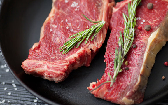 Raw fresh ribeye steak with salt, seasonings, and rosemary in a frying pan wooden background