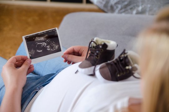 Pregnant woman expecting newborn and holding ultrasound photo and small baby shoes