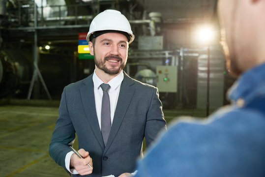 Waist up portrait of smiling bearded businessman wearing hardhat talking to worker while visiting modern plant, copy space