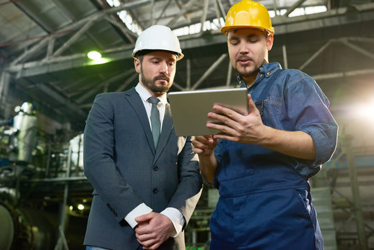 Portrait of young factory worker using digital tablet showing something to businessman wearing hardhat in workshop of modern plant, copy space
