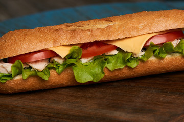 Fresh submarine sandwich with chicken fillet, cheese, fresh tomatoes, lettuce, cucumbers and onions on wooden background