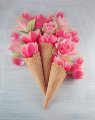 Flatlay waffle sweet ice cream cone with pink tulips blossom flowers over white wood background, top view. Spring or summer mood concept.
