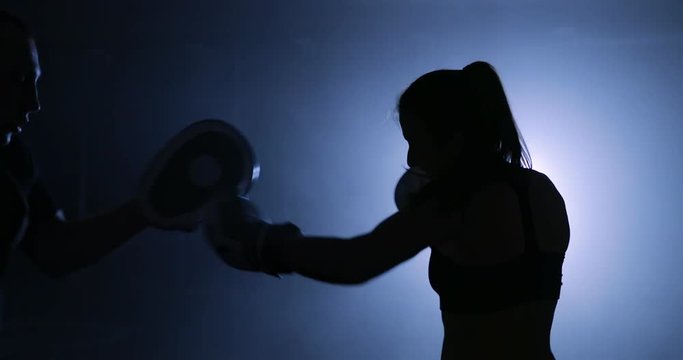 Silhouettes of a woman boxer trains her punches on a punching bag that her partner in dark gym