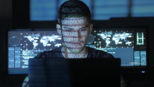 Male hacker programmer working at computer while blue code characters reflect on his face in cyber security center filled with display screens.