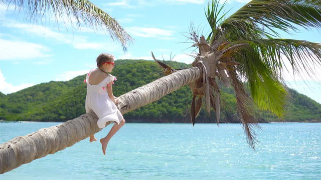 Little girl at tropical beach sitting on palm tree and havinf a lot of fun. Kid on caribbean vacation in Antigua island